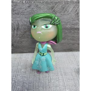 May include: A figurine of the character Disgust from the animated film "Inside Out." The figure has green skin and hair, wearing a teal dress with white flower accents, a pink scarf, and a green belt. The figure is approximately 4 inches tall.