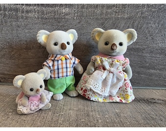 May include: Three koala bear figurines. One koala wears a plaid shirt and green pants. Another koala wears a floral dress with a white apron. The smallest koala wears a pink dress with lace trim.