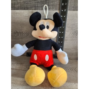 May include: A plush Mickey Mouse toy with black fur, red shorts with white circles, and yellow shoes. The toy has a loop on its head for hanging.