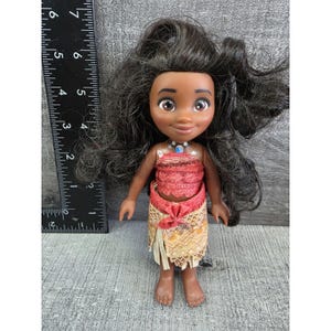 Op de afbeelding: Pop met een donkere huid, zwart haar en bruine ogen. De pop draagt een rode top, een beige rok met een rode strik en een blauwe ketting. De pop is ongeveer 15 cm hoog.