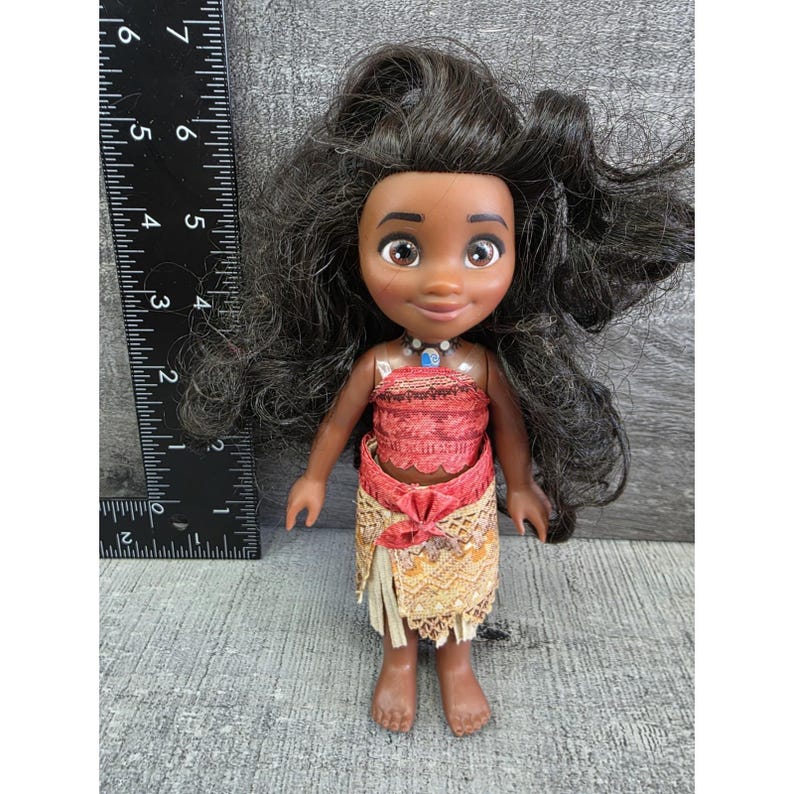 Op de afbeelding: Pop met een donkere huid, zwart haar en bruine ogen. De pop draagt een rode top, een beige rok met een rode strik en een blauwe ketting. De pop is ongeveer 15 cm hoog.