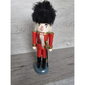 Puede incluir: Una figura de cascanueces de madera con un uniforme rojo y dorado, un sombrero de piel negra y una barba blanca. El cascanueces sostiene una espada y está de pie sobre una base azul.