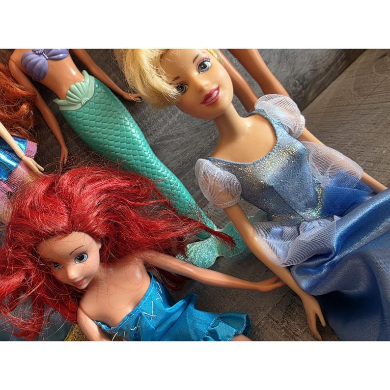 Op de afbeelding: Een verzameling poppen met personages met lang haar en kleurrijke outfits. E&eacute;n pop heeft rood haar en een blauwe jurk, een andere heeft blond haar en een blauwe jurk, en een derde pop heeft een groene zeemeerminstaart.