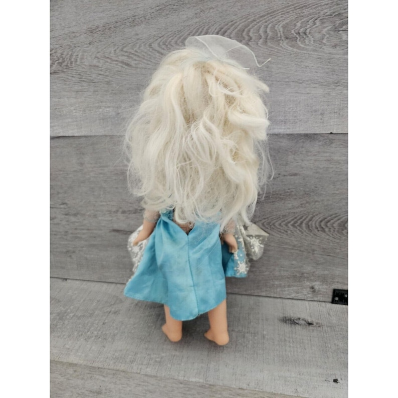 May include: A doll with long, white, curly hair wearing a light blue dress with a white snowflake pattern. The doll is standing on a wooden surface.