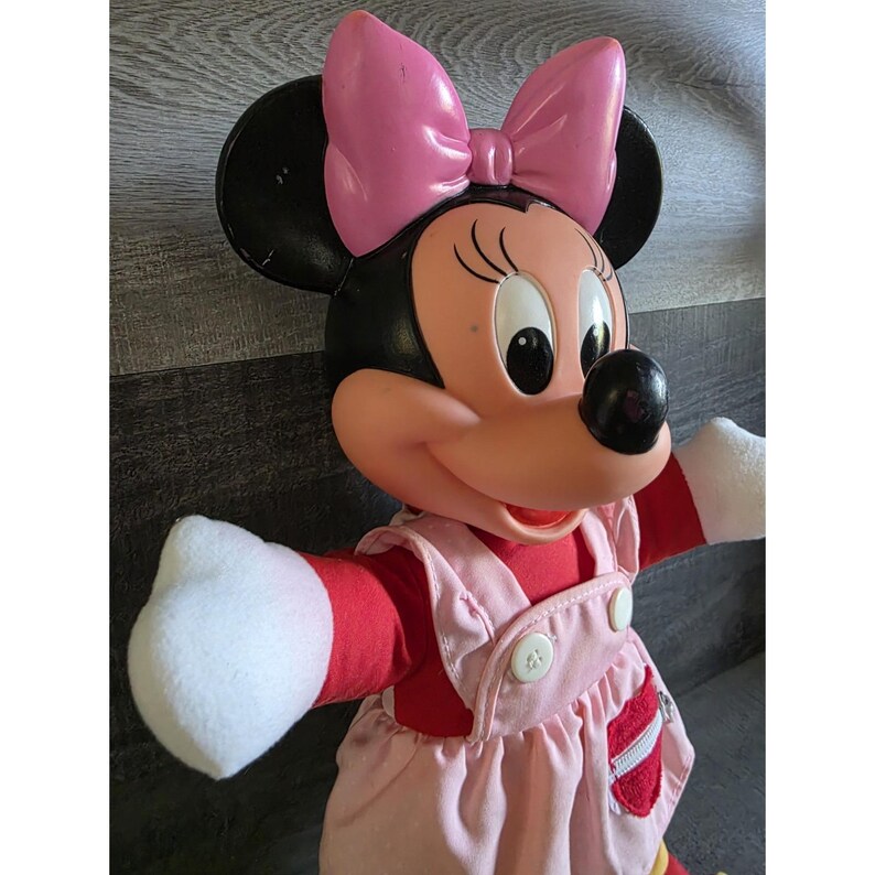 May include: A Minnie Mouse doll with a large pink bow, black ears, and a red and pink outfit. The doll has white gloves and a black nose. The doll's outfit includes a red top and pink overalls with white buttons.