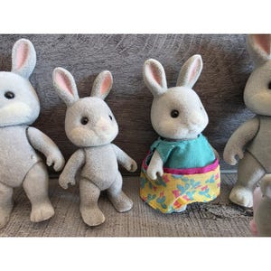 Puede incluir: Cuatro figuras de conejos grises con orejas forradas de rosa y ojos negros. Un conejo lleva una camiseta turquesa y una falda amarilla con estampado floral. Los conejos est&aacute;n sobre una superficie gris.