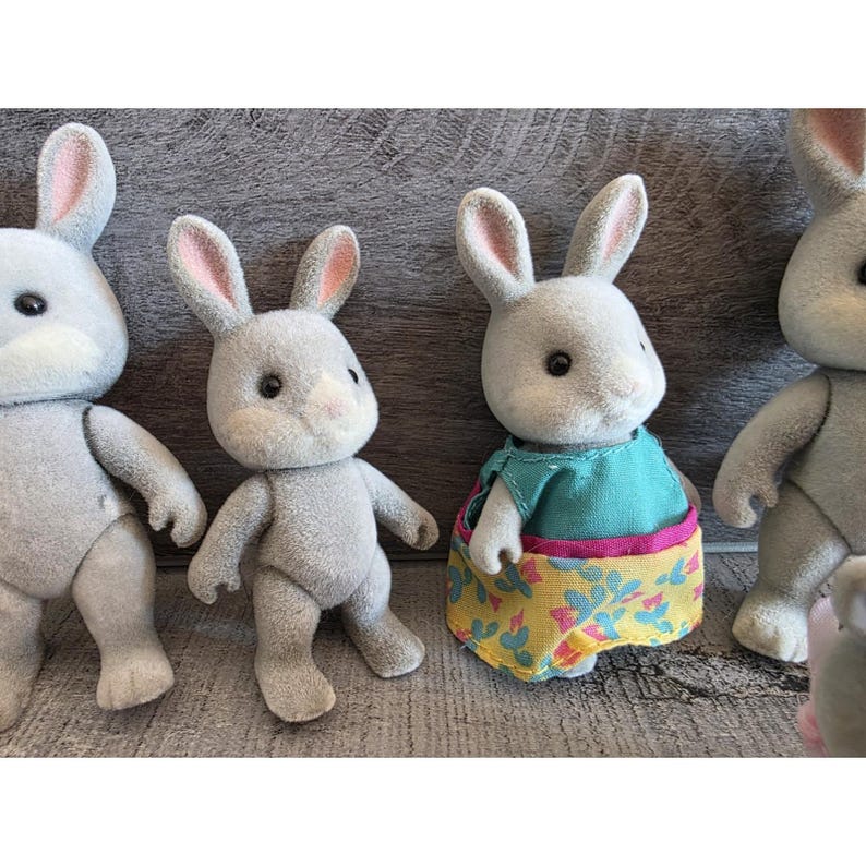 Puede incluir: Cuatro figuras de conejos grises con orejas forradas de rosa y ojos negros. Un conejo lleva una camiseta turquesa y una falda amarilla con estampado floral. Los conejos est&aacute;n sobre una superficie gris.