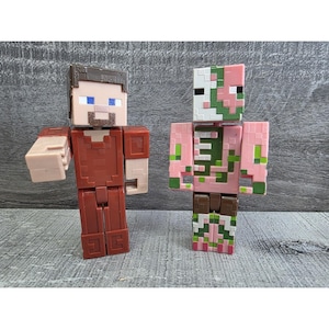 May include: Two Minecraft action figures. One figure is a brown-haired character in a red outfit with a tan arm. The other figure is a pink and green zombie-like character with a white mask. Both figures are standing on a gray surface.