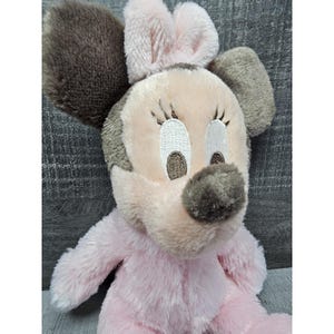 May include: A plush Minnie Mouse doll in soft pink and gray. The doll features embroidered facial details, including eyelashes and a nose. The ears are a darker gray, and the bow is a lighter pink. The doll is made of a soft, fuzzy material.
