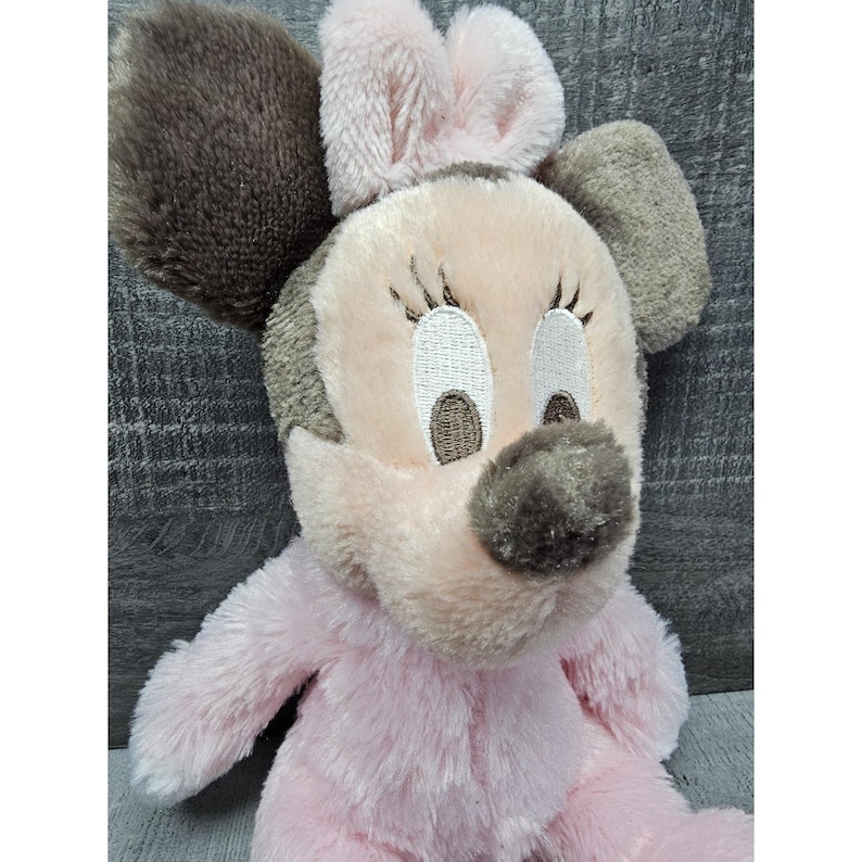 May include: A plush Minnie Mouse doll in soft pink and gray. The doll features embroidered facial details, including eyelashes and a nose. The ears are a darker gray, and the bow is a lighter pink. The doll is made of a soft, fuzzy material.