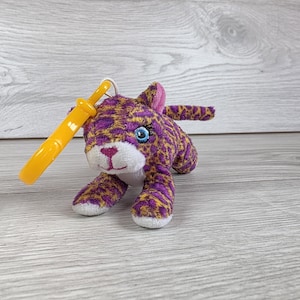 May include: A plush leopard toy featuring a purple and gold spotted pattern, bright blue embroidered eyes, and a white muzzle with a pink stitched nose. A yellow plastic clip is attached to the top of the head, allowing it to function as a keychain.
