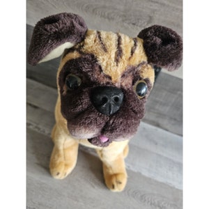 Puede incluir: Un juguete de peluche de un perro pug marrón y beige con grandes orejas marrones y ojos negros. El juguete tiene la lengua rosa que sobresale.