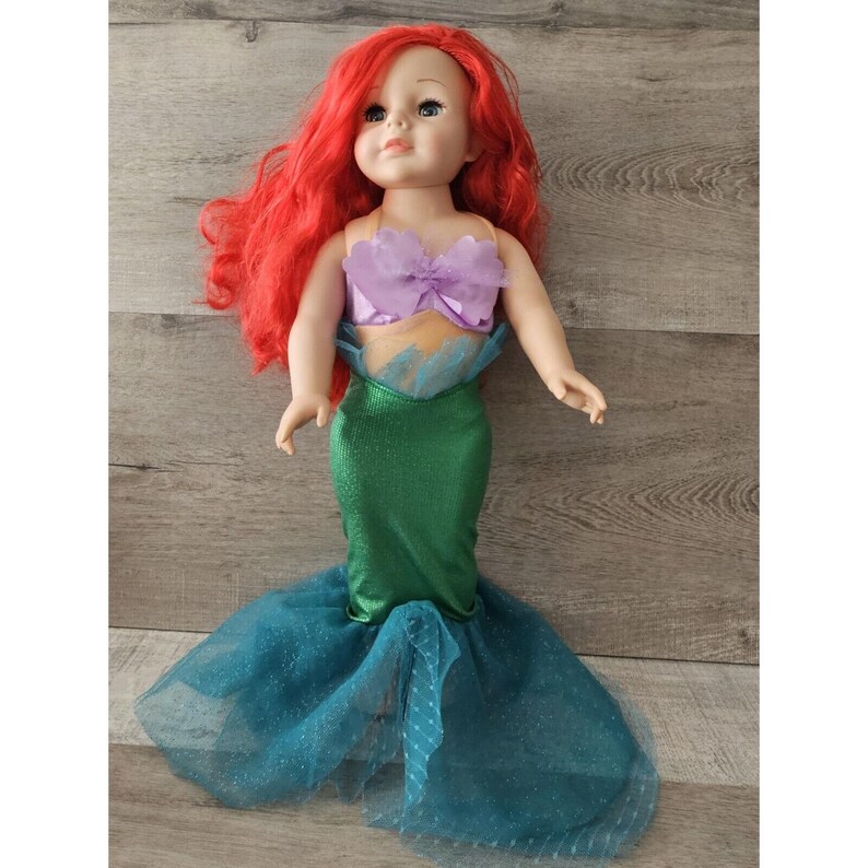 Puede incluir: Una mu&ntilde;eca de pelo rojo con un disfraz de sirena con un top morado y una cola verde con una falda de tul azul.