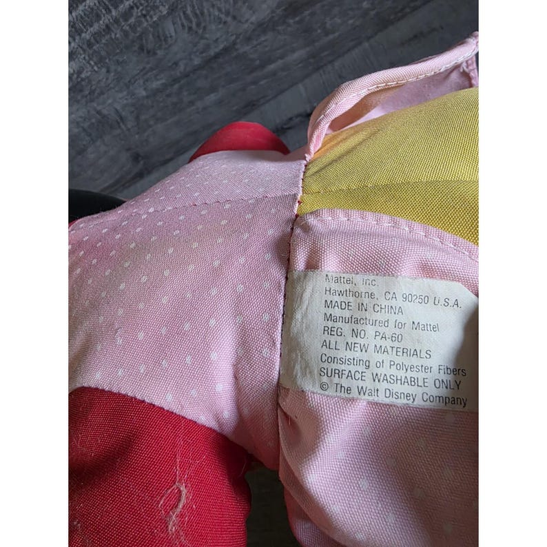 May include: Close-up of a plush toy with pink, red, and yellow fabric. The pink fabric has white polka dots. A tag reads "Mattel, Inc. Hawthorne, CA 90250 U.S.A. MADE IN CHINA Manufactured for Mattel".