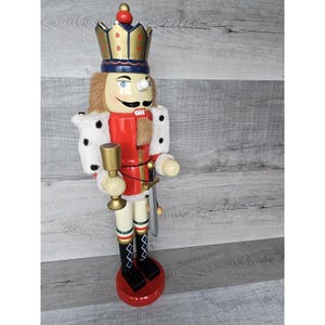 Puede incluir: Una figura de cascanueces de madera de un rey que lleva una corona dorada con detalles azules y blancos. Sostiene una copa dorada en su mano derecha y una espada en su mano izquierda. Lleva una capa roja y blanca con ribete de piel blanca.