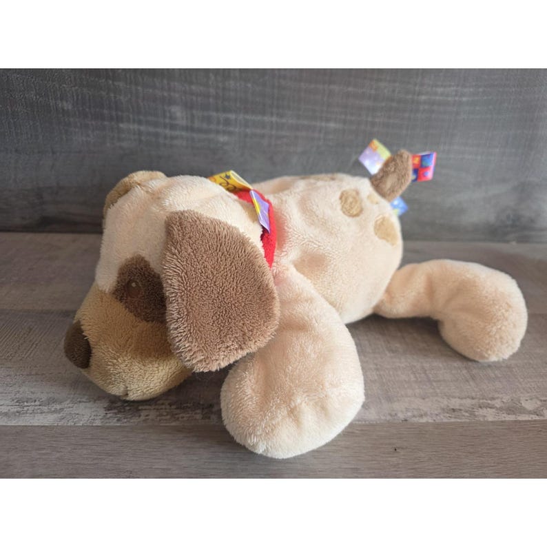 Puede incluir: Un juguete de peluche con forma de perro en tonos beige y marr&oacute;n, con orejas ca&iacute;das y una nariz cosida. El juguete tiene un collar rojo y cintas de colores. El perro est&aacute; tumbado sobre una superficie de madera.