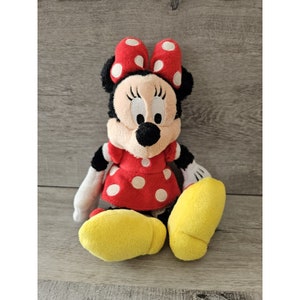 K&ouml;nnte beinhalten: Ein rotes und wei&szlig; gepunktetes Pl&uuml;schtier von Minnie Maus. Sie tr&auml;gt ein rotes Kleid mit wei&szlig;en Punkten und eine rote Schleife mit wei&szlig;en Punkten. Sie hat schwarze Augen und eine schwarze Nase. Sie sitzt mit gekreuzten Beinen und ihren Armen an den Seiten.