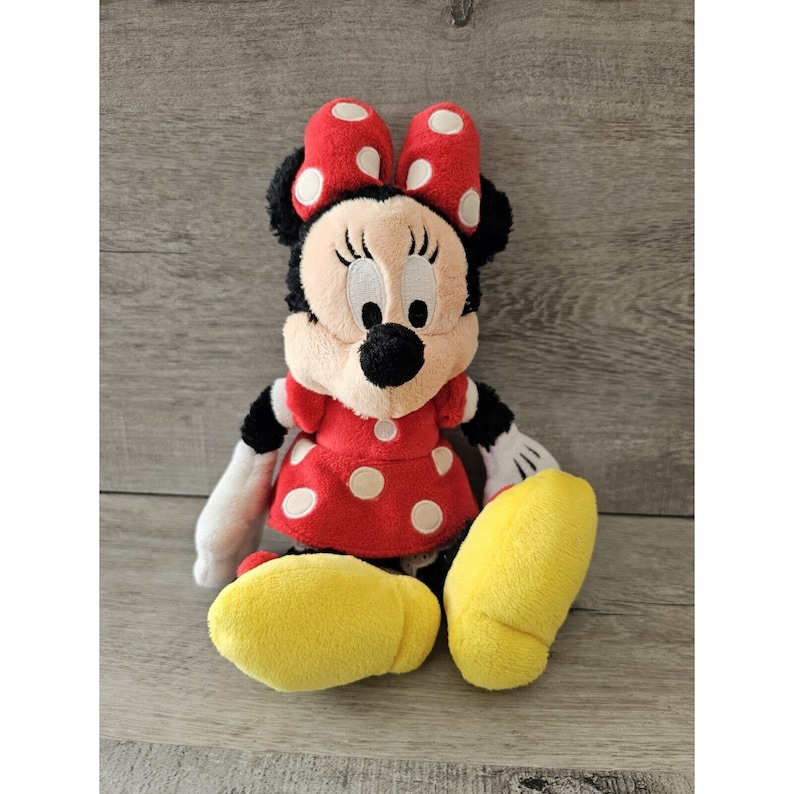 K&ouml;nnte beinhalten: Ein rotes und wei&szlig; gepunktetes Pl&uuml;schtier von Minnie Maus. Sie tr&auml;gt ein rotes Kleid mit wei&szlig;en Punkten und eine rote Schleife mit wei&szlig;en Punkten. Sie hat schwarze Augen und eine schwarze Nase. Sie sitzt mit gekreuzten Beinen und ihren Armen an den Seiten.