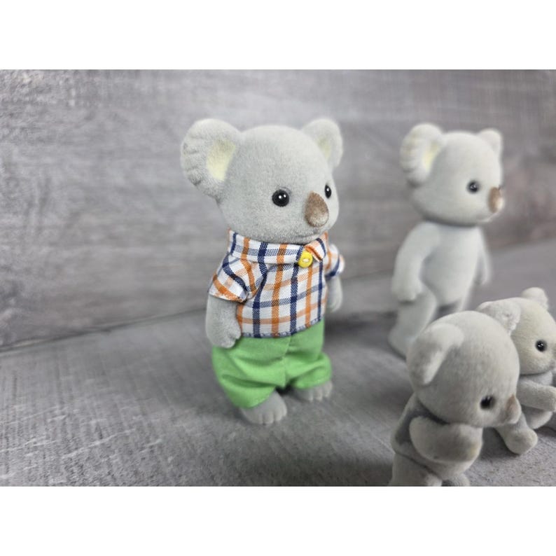 May include: A group of gray koala figurines. One koala wears a plaid shirt with orange, blue, and white squares, and green pants. Other koalas are visible in the background. The figurines have black eyes and tan noses.