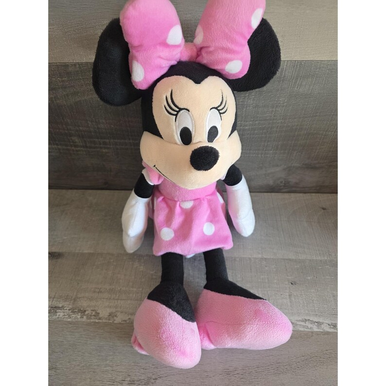 May include: A plush Minnie Mouse doll. The doll has black ears, a pink bow with white polka dots, and a pink dress with white polka dots. The doll's feet and hands are pink and white, respectively. The doll is standing on a wooden surface.