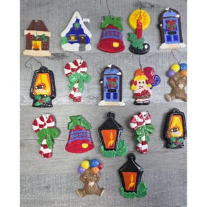 May include: A selection of Christmas tree decorations in diverse shapes, such as a house, bell, candle, and lantern. The ornaments are brightly coloured and feature festive details like candy canes and a teddy bear with balloons. Each decoration has a small loop for hanging.