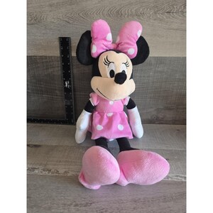 May include: A plush Minnie Mouse doll, featuring black ears, a beige face, and a pink dress with white polka dots. The doll has a matching pink bow with white polka dots and large pink feet. A black ruler is next to the doll.