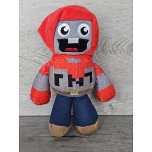 Może przedstawiać: Pluszowa zabawka w kolorze szarym i czerwonym z szerokim, zębatym uśmiechem, ubrana w czerwoną kurtkę z kapturem i niebieskie dżinsy. Zabawka ma napis "TNT" na klatce piersiowej.