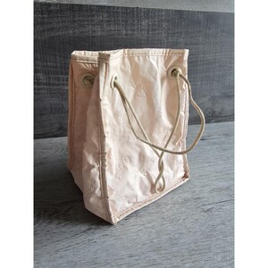 May include: A light pink tote bag with a drawstring closure and beige rope handles. The bag has a repeating embossed pattern and is made of a shiny, possibly satin-like material. The bag is rectangular and stands upright.
