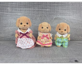 May include: Three miniature dog figurines. The dogs have tan fur and black eyes. One dog wears a pink dress with a white apron, another a pink floral dress, and the third a plaid outfit.