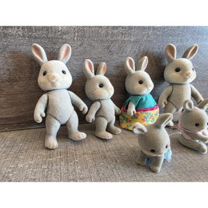 Puede incluir: Colecci&oacute;n de seis figuras de conejos grises con caras blancas y orejas forradas en rosa. Un conejo lleva una blusa turquesa y una falda floral. Otro tiene una cinta rosa. Los conejos est&aacute;n sobre una superficie de madera gris.