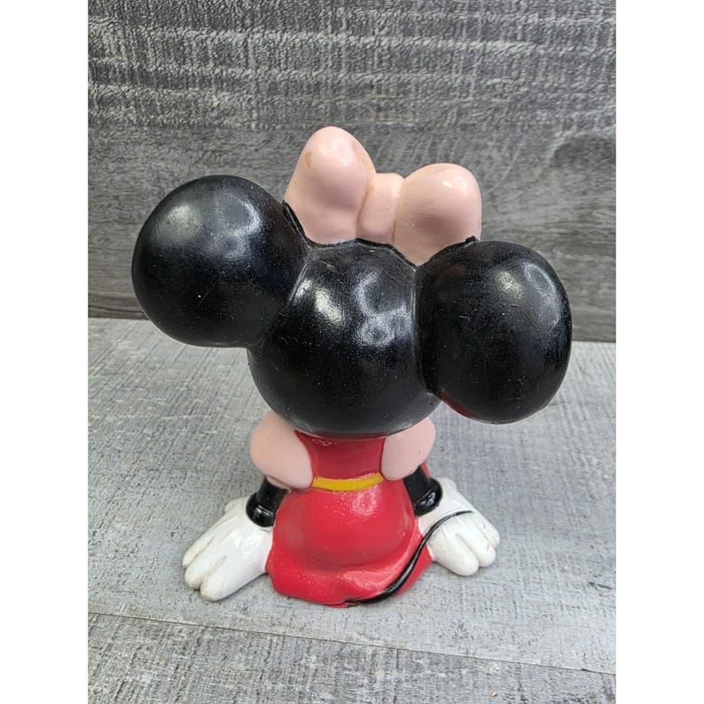 May include: A Minnie Mouse figurine, featuring a black head with large ears and a pink bow. The figurine is wearing a red dress with a yellow belt and white gloves. The figure is in a seated position.