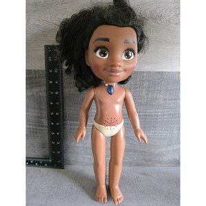 May include: A doll with dark skin, black hair, and large brown eyes. The doll wears a white diaper and a blue pendant necklace. A ruler is placed next to the doll, showing measurements in inches.