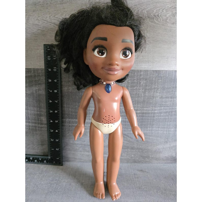 May include: A doll with dark skin, black hair, and large brown eyes. The doll wears a white diaper and a blue pendant necklace. A ruler is placed next to the doll, showing measurements in inches.