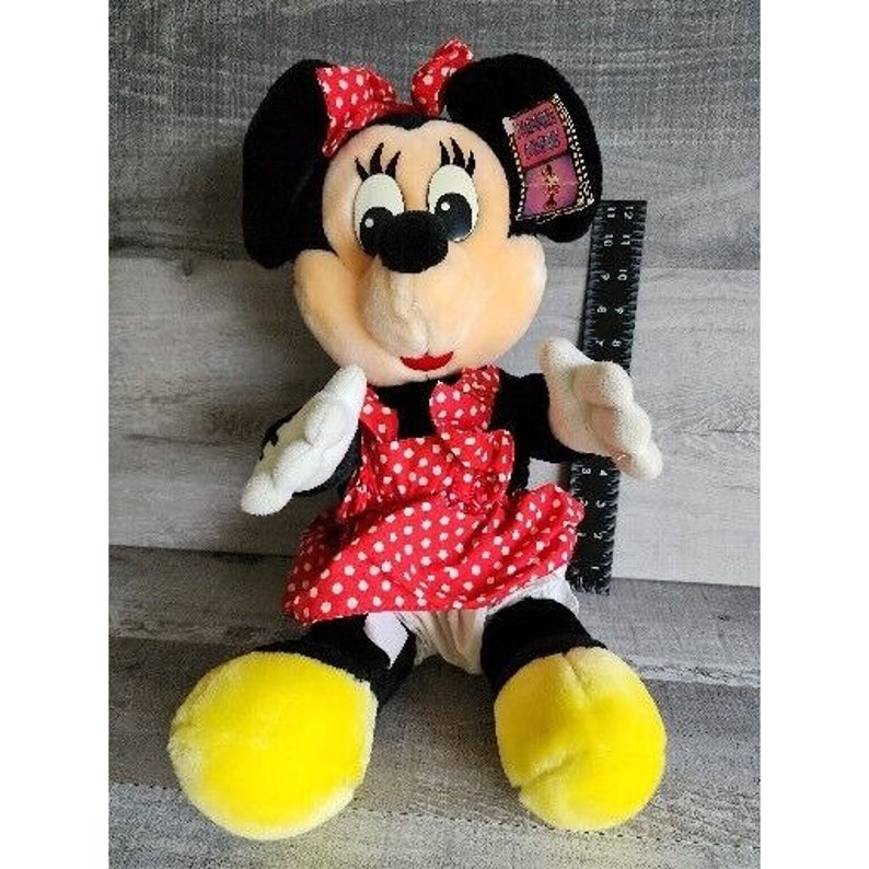 Pu&ograve; includere: Un giocattolo di peluche di Minnie Mouse che indossa un vestito a pois rossi e bianchi. Ha un fiocco nero nei capelli e un'etichetta gialla che dice "Disney's Friends Forever".