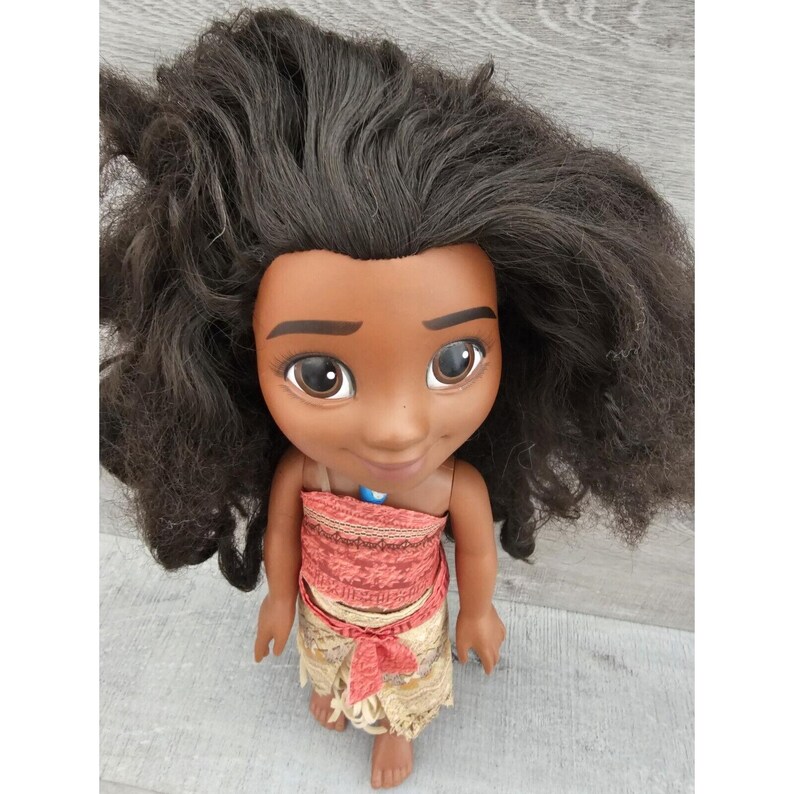 Puede incluir: Una mu&ntilde;eca de piel morena con el pelo largo y negro rizado. Lleva un top estampado en rojo y marr&oacute;n y una falda beige con una faja marr&oacute;n. La mu&ntilde;eca sonr&iacute;e y tiene ojos marrones.