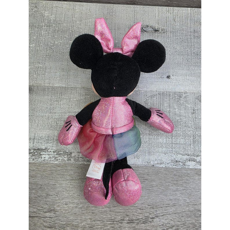 K&ouml;nnte beinhalten: Ein Pl&uuml;sch Minnie Mouse Puppe mit schwarzen Ohren, einer rosa Schleife und einem rosa Outfit. Die Puppe hat eine rosa Jacke, rosa Handschuhe und rosa Schuhe. Der Rock ist ein Regenbogen aus Farben. Die Puppe ist ein Disney-Produkt.