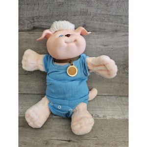 May include: A pink plush dog with white fur on its head, wearing a blue shirt and a brown collar with a gold tag. The dog has blue eyes and a happy expression.
