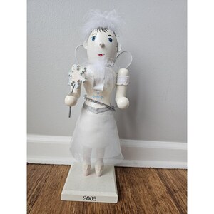 May include: White porcelain nutcracker figurine of a fairy with a snowflake wand, wearing a white dress and a white feathered crown. The base of the nutcracker is white and has the year 2005 printed on it.