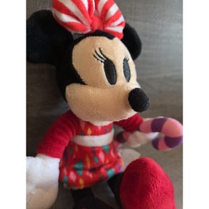 May include: A plush Minnie Mouse doll with a red dress featuring a colorful diamond pattern. She has a red and white striped bow, black ears, and a black nose. Minnie is holding a candy cane.
