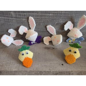 May include: Handmade Easter decorations featuring white and pink bunnies and yellow and orange carrots. The bunnies have white bodies, pink ears, and some have purple or blue accents. The carrots have green tops and orange bodies.