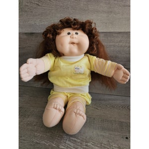 May include: A vintage doll with a round face, brown curly hair, and large eyes. The doll wears a yellow outfit with a white cat graphic on the chest. The outfit includes a long-sleeved shirt and shorts with pink and white stripes.