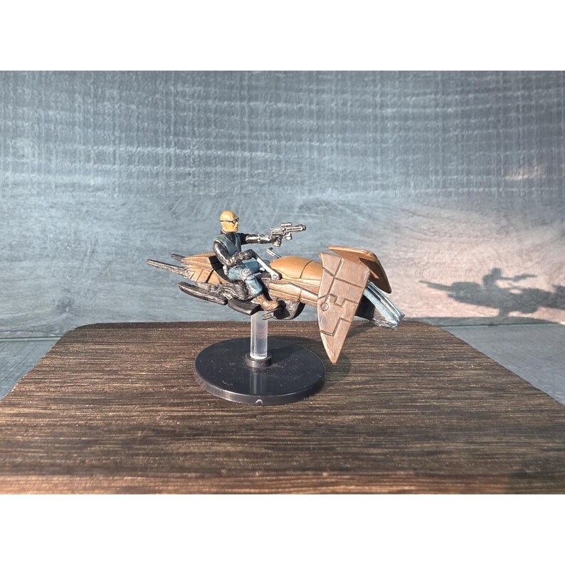 May include: A miniature toy of a futuristic vehicle with a seated figure. The vehicle is primarily bronze with black and silver accents. The figure is wearing a yellow helmet and dark clothing, holding a weapon. The toy is mounted on a clear stand.
