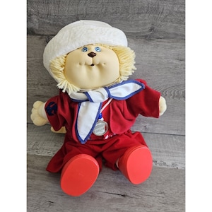 May include: A plush toy with a white sailor hat, blonde hair, and a red and blue sailor outfit. The toy is wearing a silver medal with the words "Little  Sailor" on it.