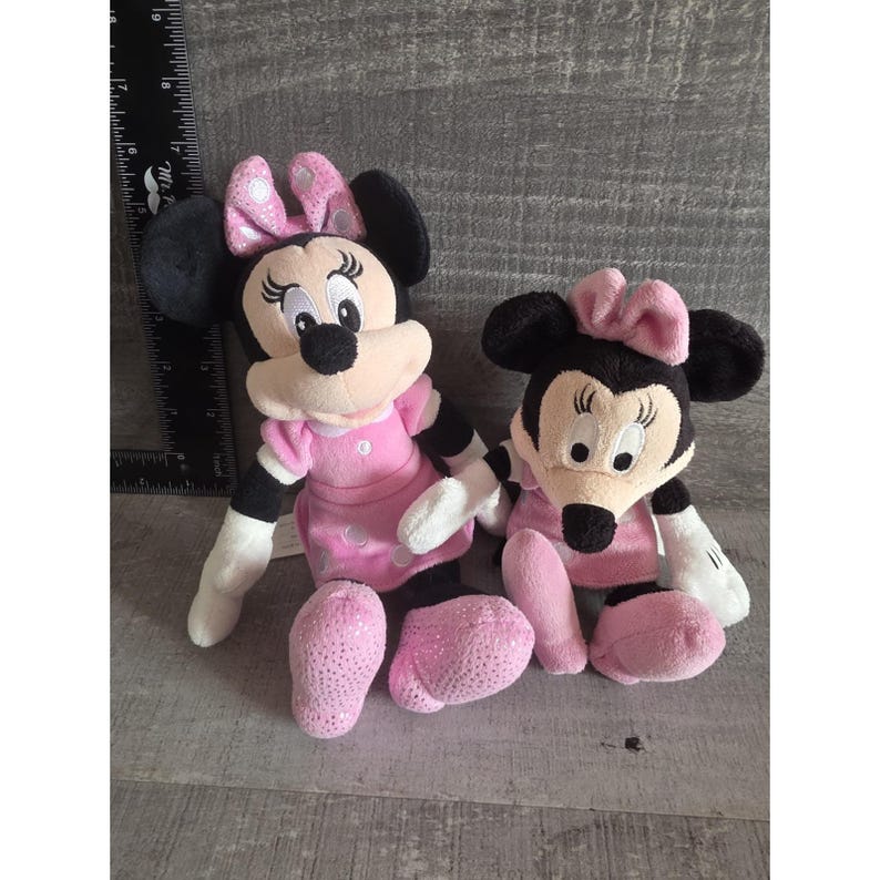 May include: Two Minnie Mouse plush toys. The toys have black ears, a pink bow, and a pink dress. One toy has sequined pink shoes. The toys are sitting on a gray surface.