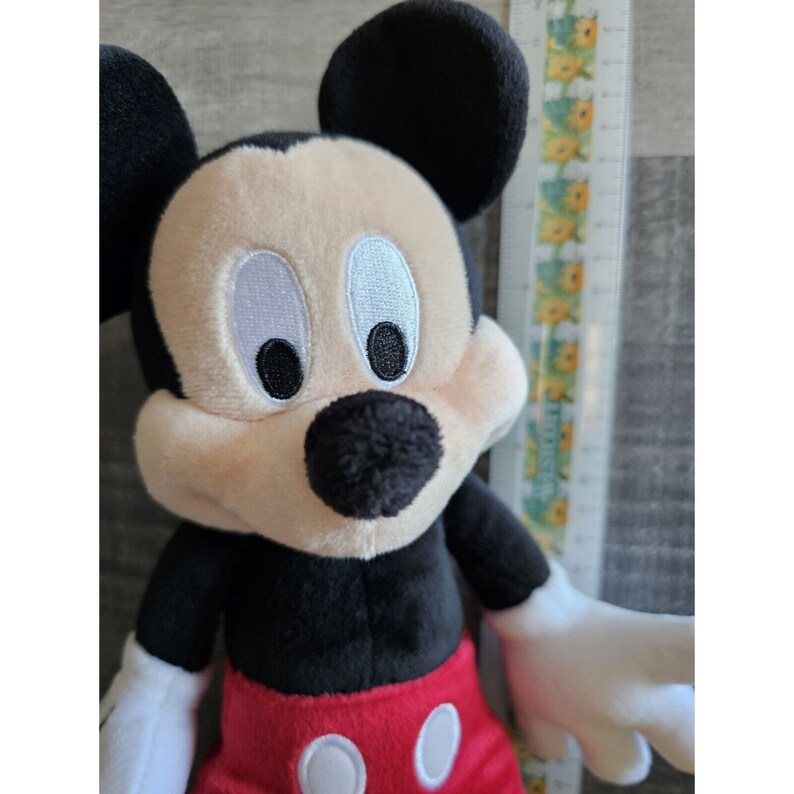 Puede incluir: Un juguete de peluche de Mickey Mouse con orejas negras, una nariz negra y pantalones cortos rojos con botones blancos. El juguete tiene una cara beige con ojos negros bordados y un contorno blanco.