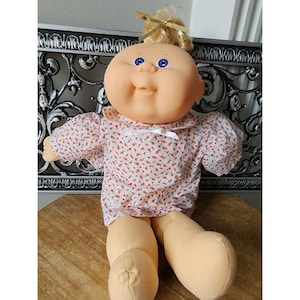 Può includere: Una bambola Cabbage Patch Kid con capelli biondi e occhi azzurri che indossa un vestito bianco con stampa floreale rossa e blu.