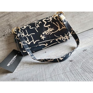 May include: A black handbag with a cream-coloured abstract design and a matching strap. The bag has gold-coloured rings and a tag that reads PRETTYLITTLETHING. The bag is on a wooden surface.