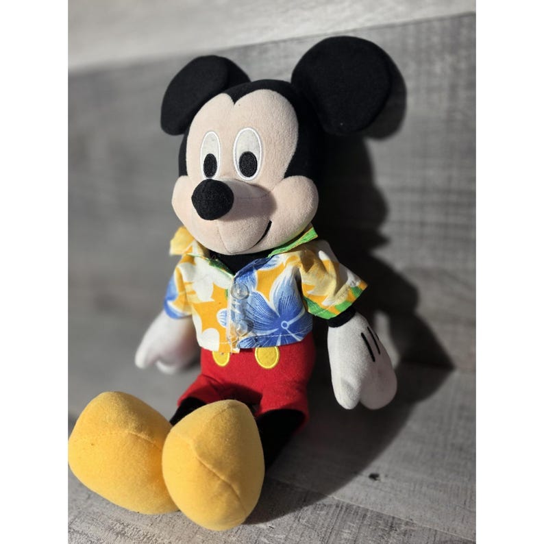 Puede incluir: Mu&ntilde;eco de peluche de Mickey Mouse con una camisa hawaiana con estampados florales azules, detalles amarillos y verdes, pantalones cortos rojos y zapatos amarillos. El mu&ntilde;eco tiene orejas negras, nariz negra y guantes blancos. El mu&ntilde;eco est&aacute; sentado.
