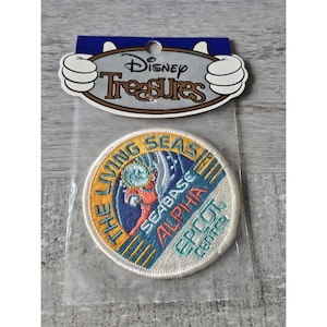 May include: A white, round embroidered patch with the text "The Living Seas Seabase Alpha Epcot Center" in a blue, yellow, and orange colour scheme. The patch features a stylised image of a diver in a blue and orange wetsuit.