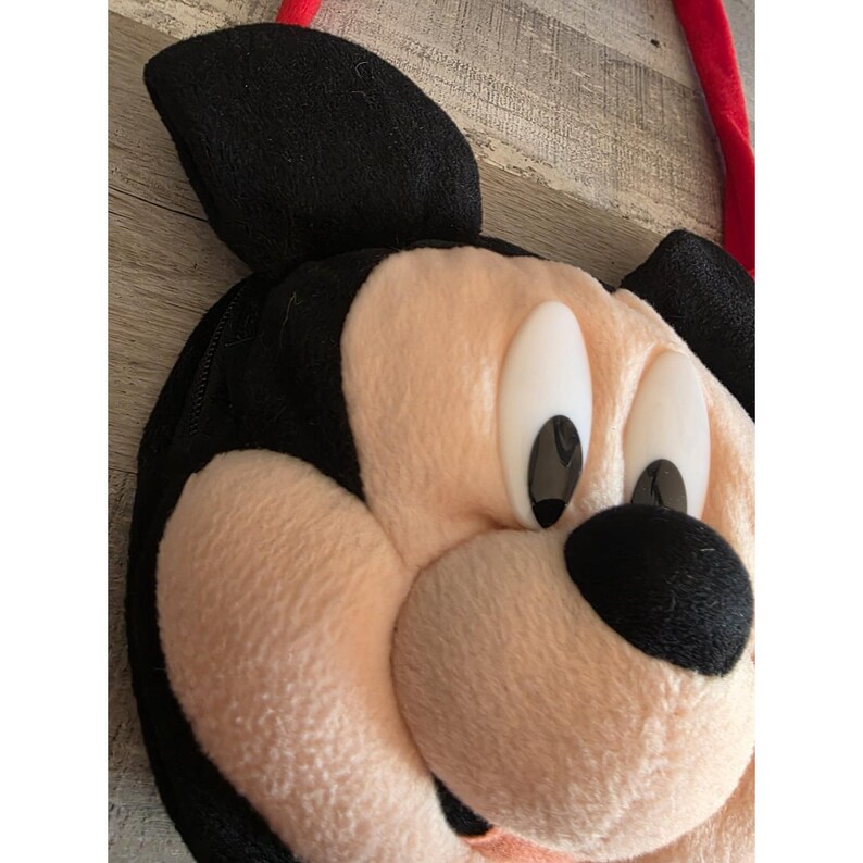 May include: A plush Mickey Mouse face bag with black ears, a black nose, and large white and black eyes. The bag has a peach-colored face and a red strap. The bag is made of soft, plush material.